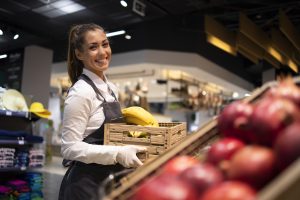 Moça sorridente, com cabelo preso e luvas, abastecendo uma prateleira de frutas em um supermercado, segurando uma caixa com bananas e olhando para a câmera.